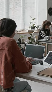 Vertical timelapse shot of busy working day of female programmer at IT startup - sitting at desk, developing computer code on laptop, testing on digital tablet, making notes, talking to colleagues
